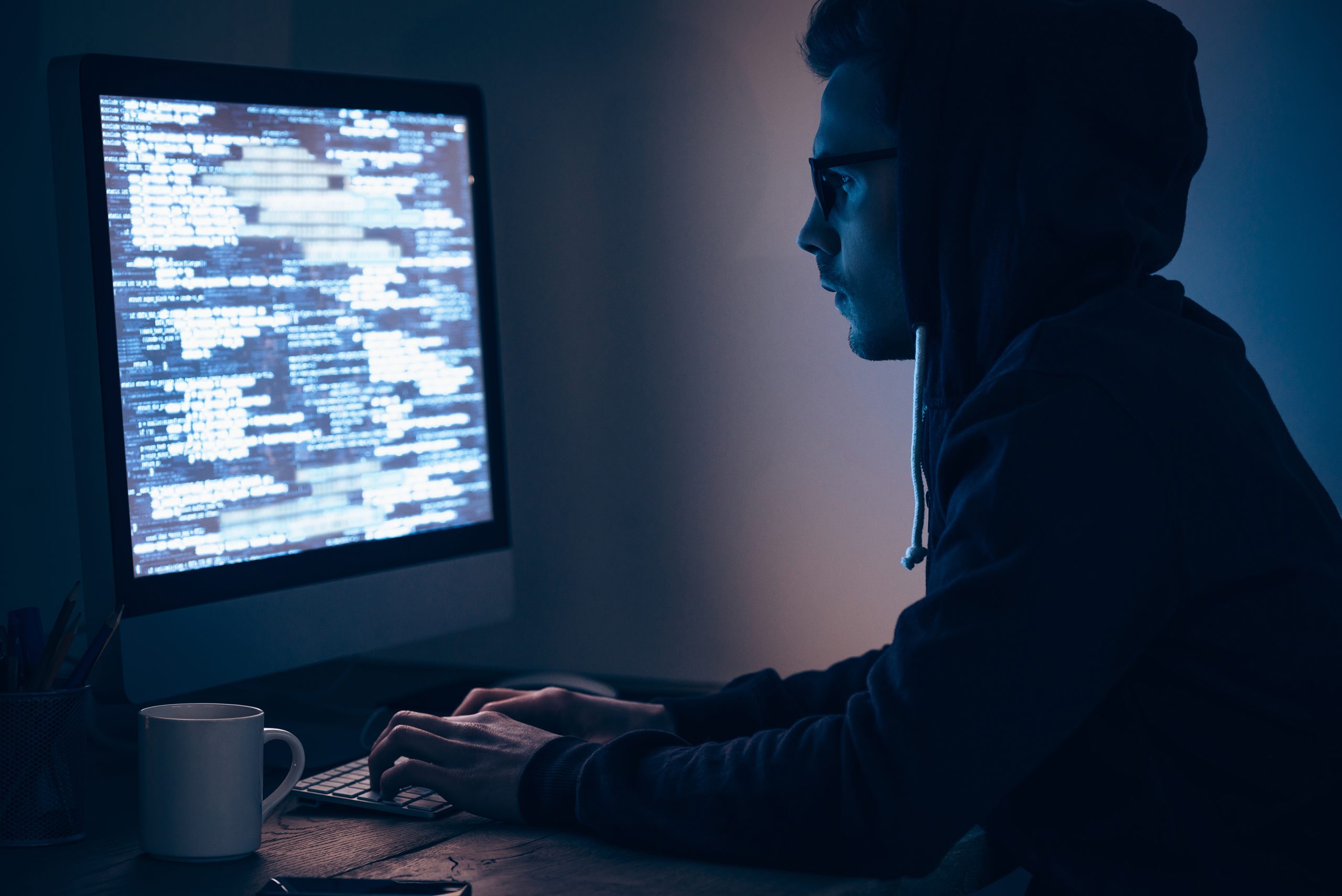 Computer spy. Side view of young man typing and looking at computer monitor while sitting at the table in dark room